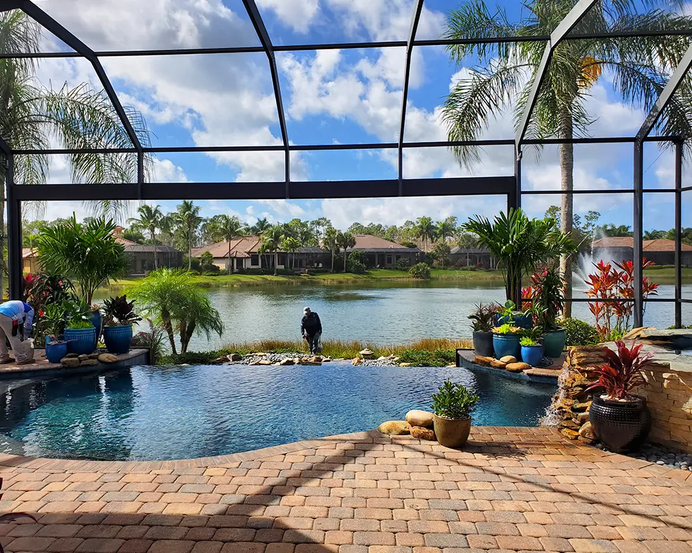 Enhancing Waterfront Properties with Custom Hardscaping. Featuring a photo of a pool deck being constructed in Southwest Florida, with a giant open window facing a retention pond, surrounded by hardscaping.
