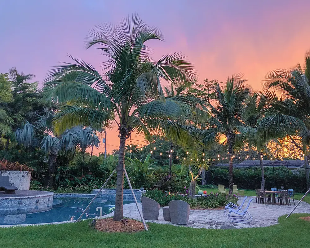 Designing Outdoor Spaces for Entertaining: Florida Style. Featuring a photo of a Florida backyard outdoor space with custom hardscaping, palm trees, and a pool.