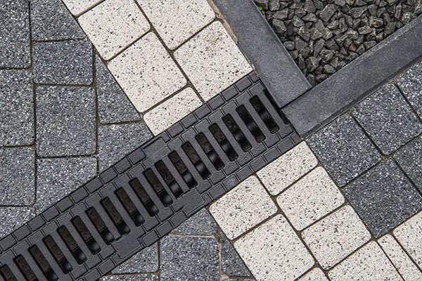 Channel drain with metal grating installed on a black and white paver hardscaped deck, feeding under a gravel surface.