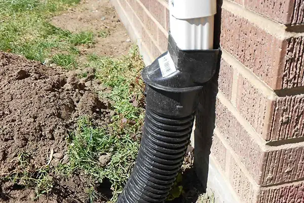Black pipe downspout extension attached to a white gutter, feeding into a trench dug into the ground below the downspout and guiding the water away from the house.